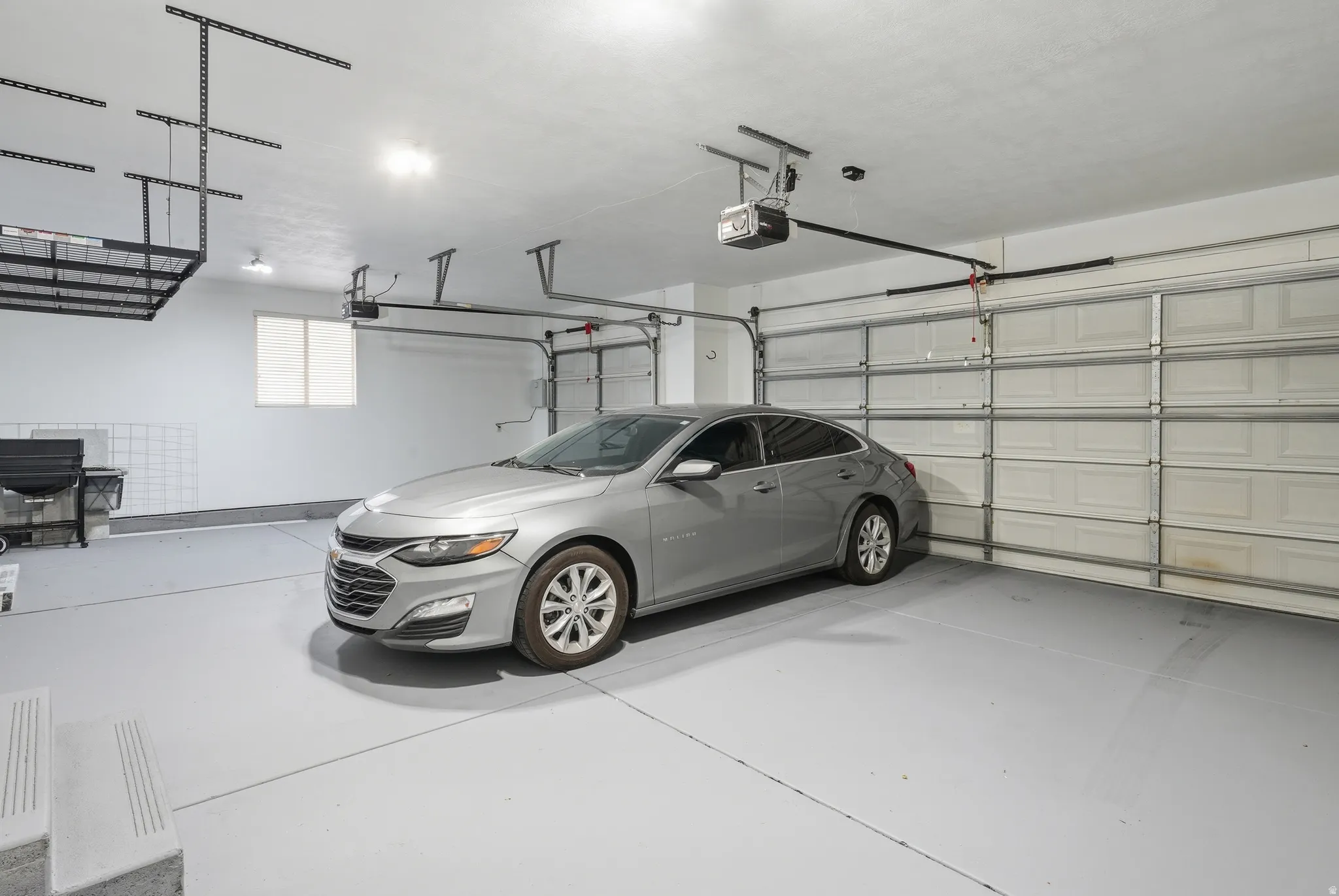Dual garage doors, overhead storage racks, light-colored flooring, window, and automated garage door openers