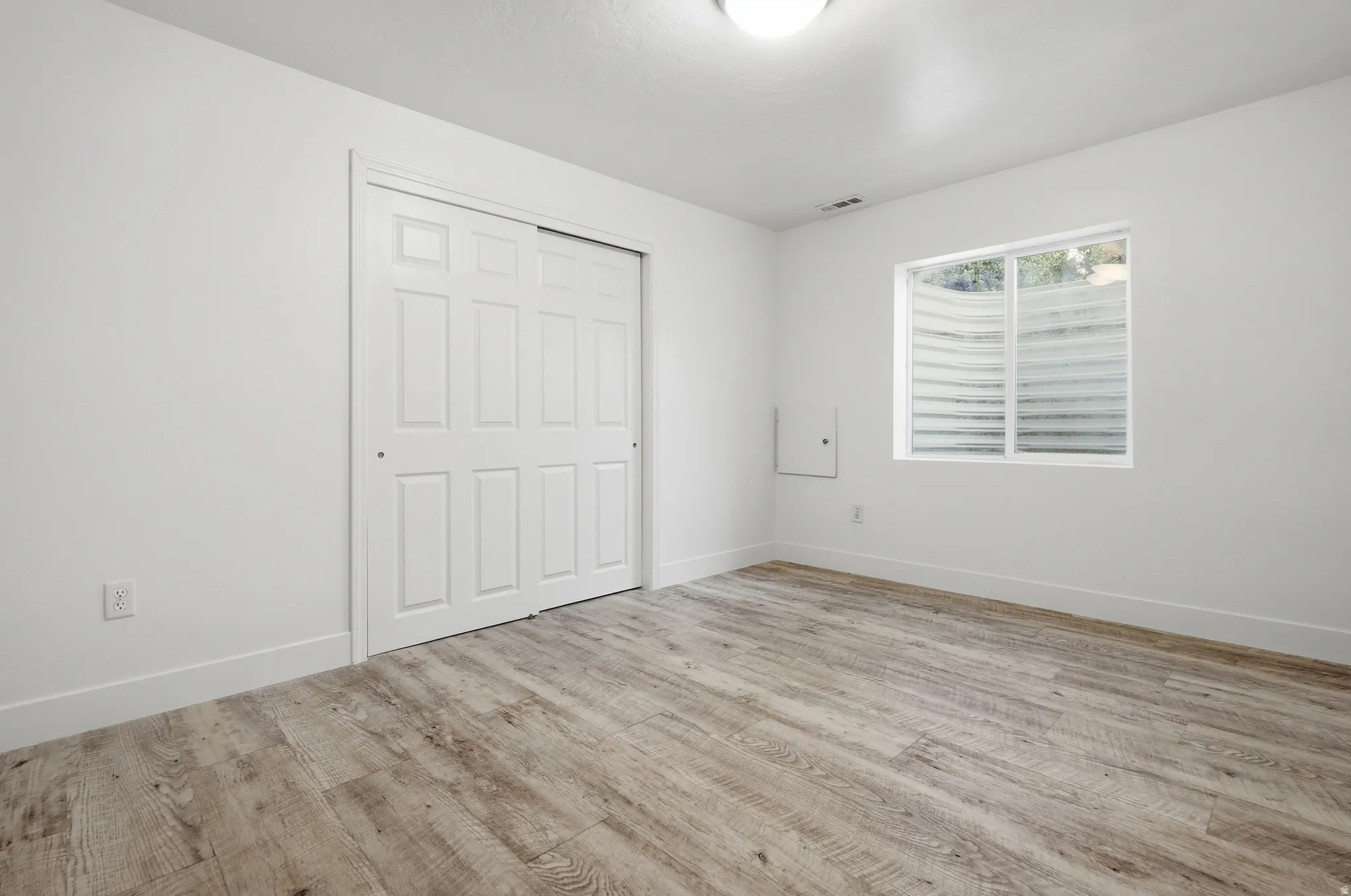 Bright bedroom showcasing wood-finish flooring, a large closet with paneled doors, and a window with plantation shutters allowing natural light throughout the space