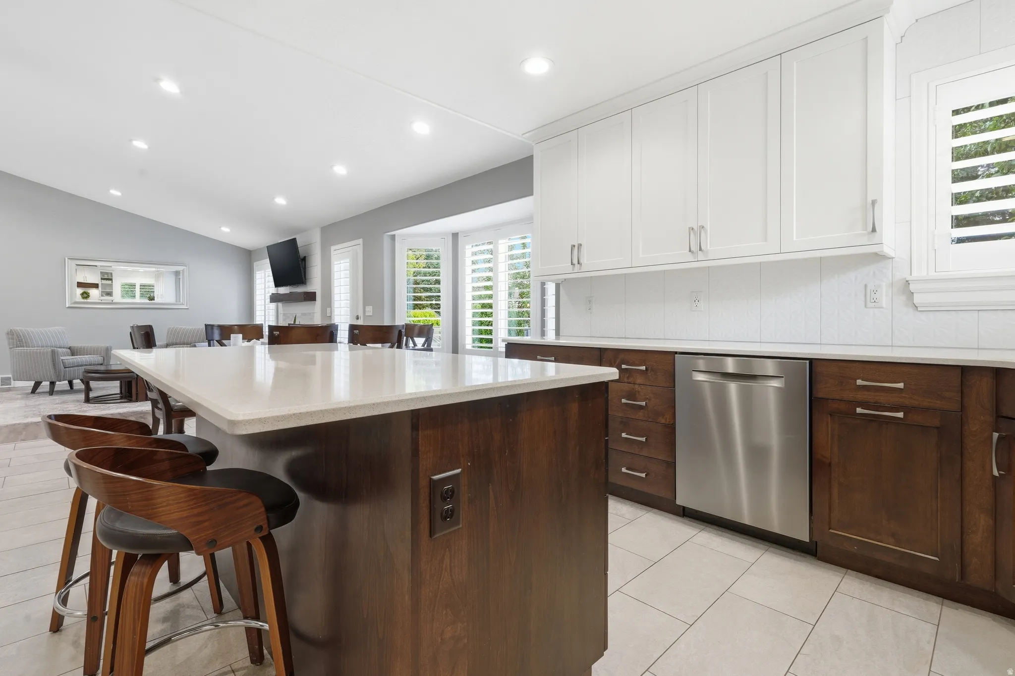 Gourmet kitchen featuring white shaker cabinetry, a two-tier island with waterfall-edge countertop, dark wood base cabinetry, stainless steel appliances, recessed and pendant lighting, tile flooring, and seamless integration with the adjoining living spac