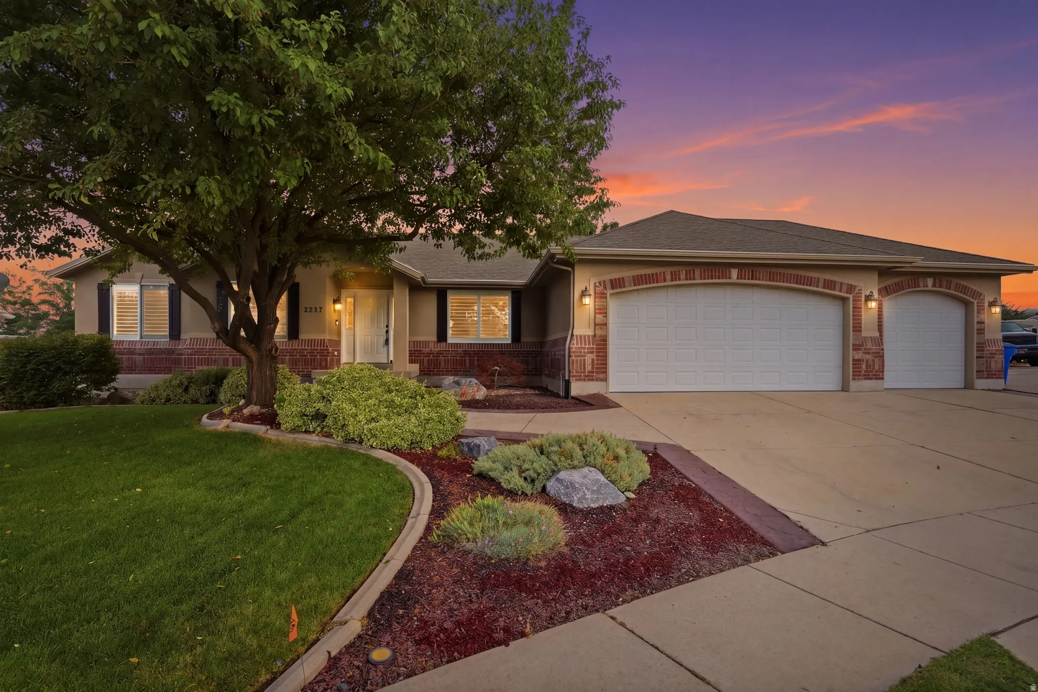 Stunning brick and stone residence showcasing a two-car garage, mature landscaping with curved hardscape borders, and professional outdoor lighting