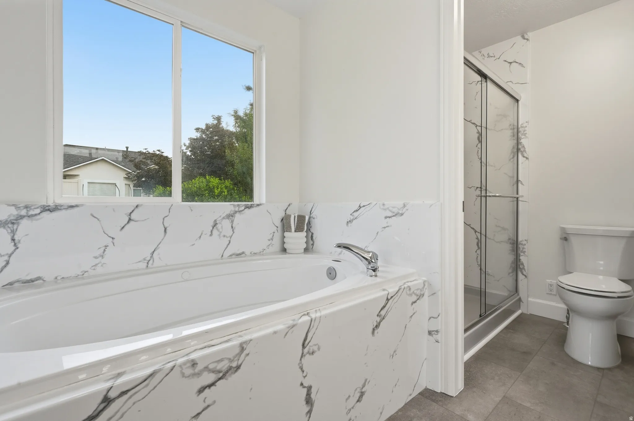 Spa-inspired primary bathroom featuring a marble-look tile surround, soaking tub with chrome fixtures, walk-in shower with frameless glass enclosure, and dual vanity
