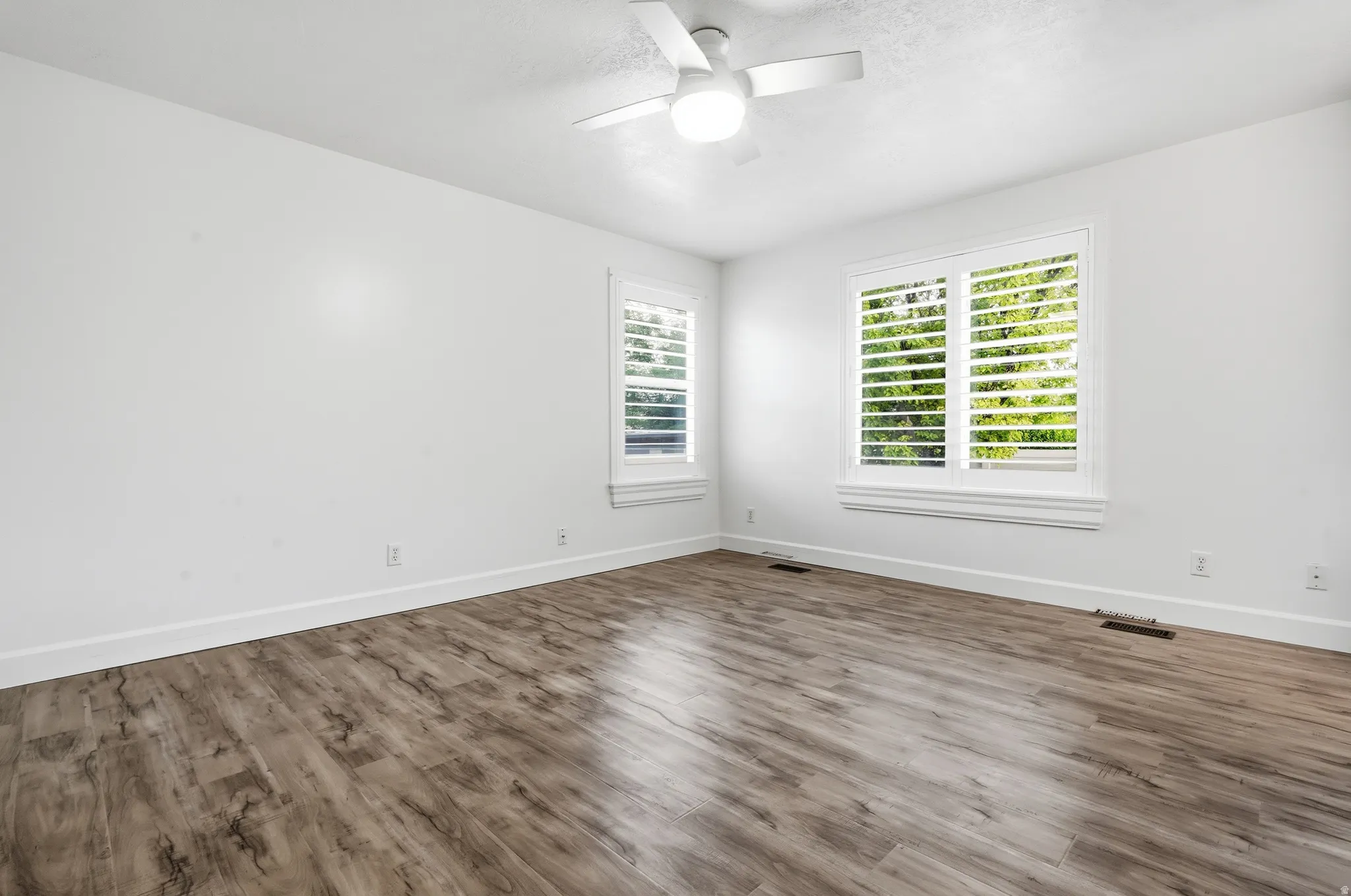 Bright, light-filled bedroom featuring wood-finish plank flooring, ceiling fan with integrated lighting, and dual windows with plantation shutters allowing natural light throughout the space