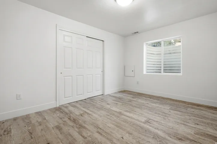 Bright bedroom showcasing wood-finish flooring, a large closet with paneled doors, and a window with plantation shutters allowing natural light throughout the space