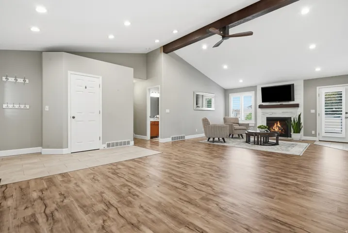 Vaulted ceiling with exposed beam, wood-finish flooring, white brick fireplace with wood mantel, recessed lighting, and white plantation shutters