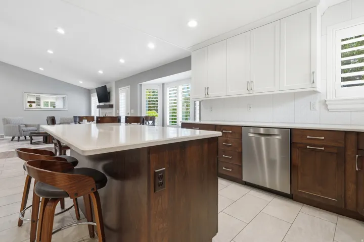 Gourmet kitchen featuring white shaker cabinetry, a two-tier island with waterfall-edge countertop, dark wood base cabinetry, stainless steel appliances, recessed and pendant lighting, tile flooring, and seamless integration with the adjoining living spac
