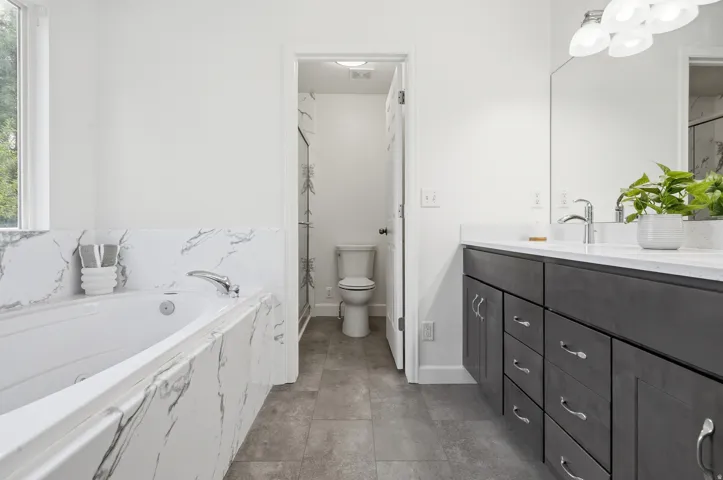 Spa-inspired primary bathroom featuring a freestanding soaking tub with marble surround, dual-sink vanity with charcoal cabinetry and chrome hardware, quartz countertops, large frameless mirror, and tile flooring throughout