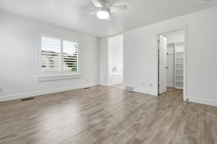 Bright and spacious bedroom featuring wood-finish flooring, large windows with plantation shutters allowing abundant natural light, ceiling fan with integrated lighting, walk-in closet with built-in shelving, and fresh white painted walls