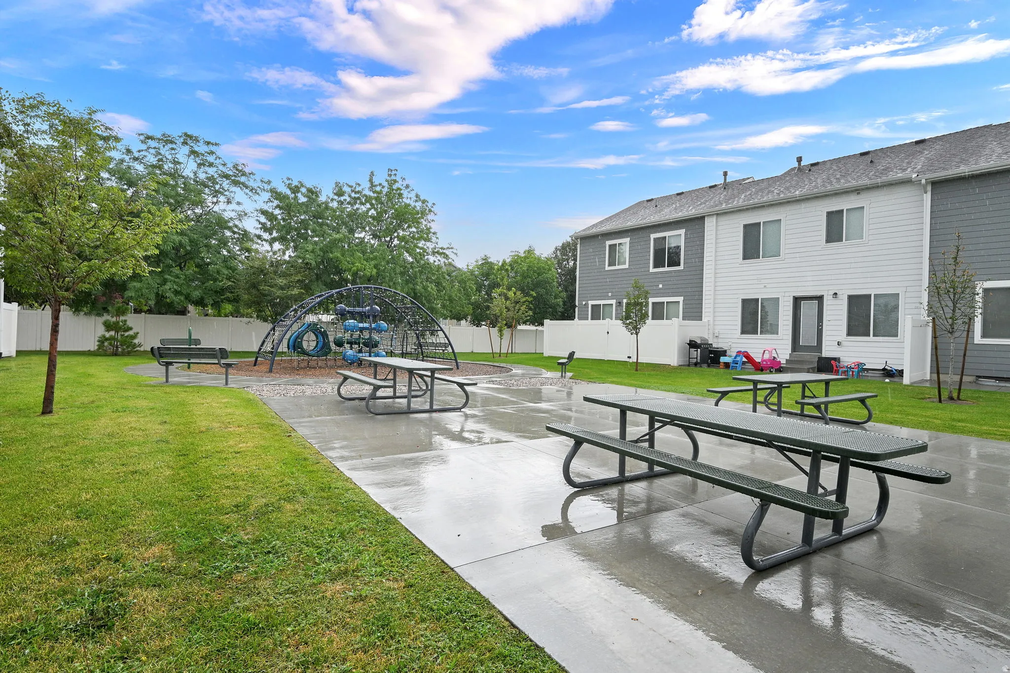 Expansive outdoor recreational area featuring a concrete patio with attached picnic tables, a multi-level playground structure, and mature landscaping with established trees