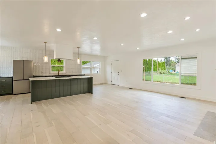 Open-concept living space featuring light wood-finish flooring throughout, a dark-finished kitchen island with vertical paneling, stainless steel appliances, recessed ceiling lighting, and expansive windows framing landscaped grounds
