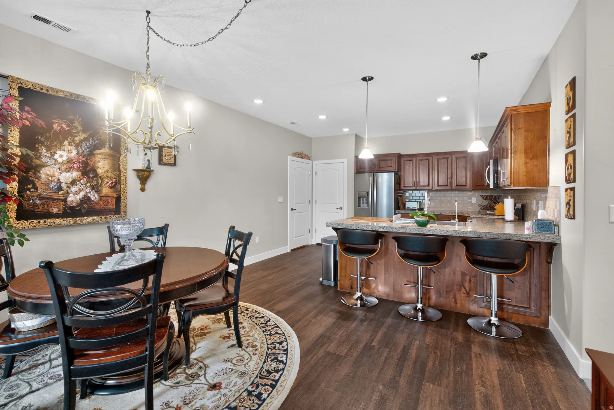 Open-concept dining and kitchen area featuring warm wood-finish flooring throughout, a round dining table with seating for four, and a chef's kitchen with rich wood cabinetry, stainless steel appliances, and a large island with seating for three