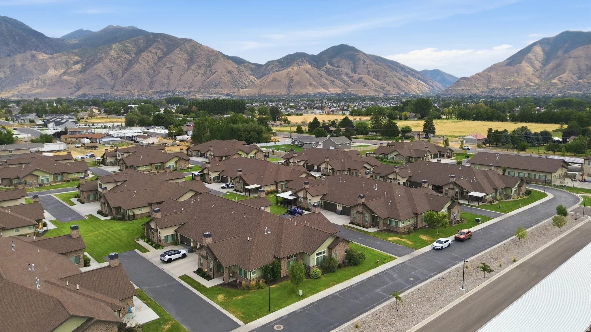 Aerial view of a newly constructed residential community featuring multiple brown-roofed townhomes arranged along curved streets with manicured lawns, mature landscaping, and mountain vistas in the distance
