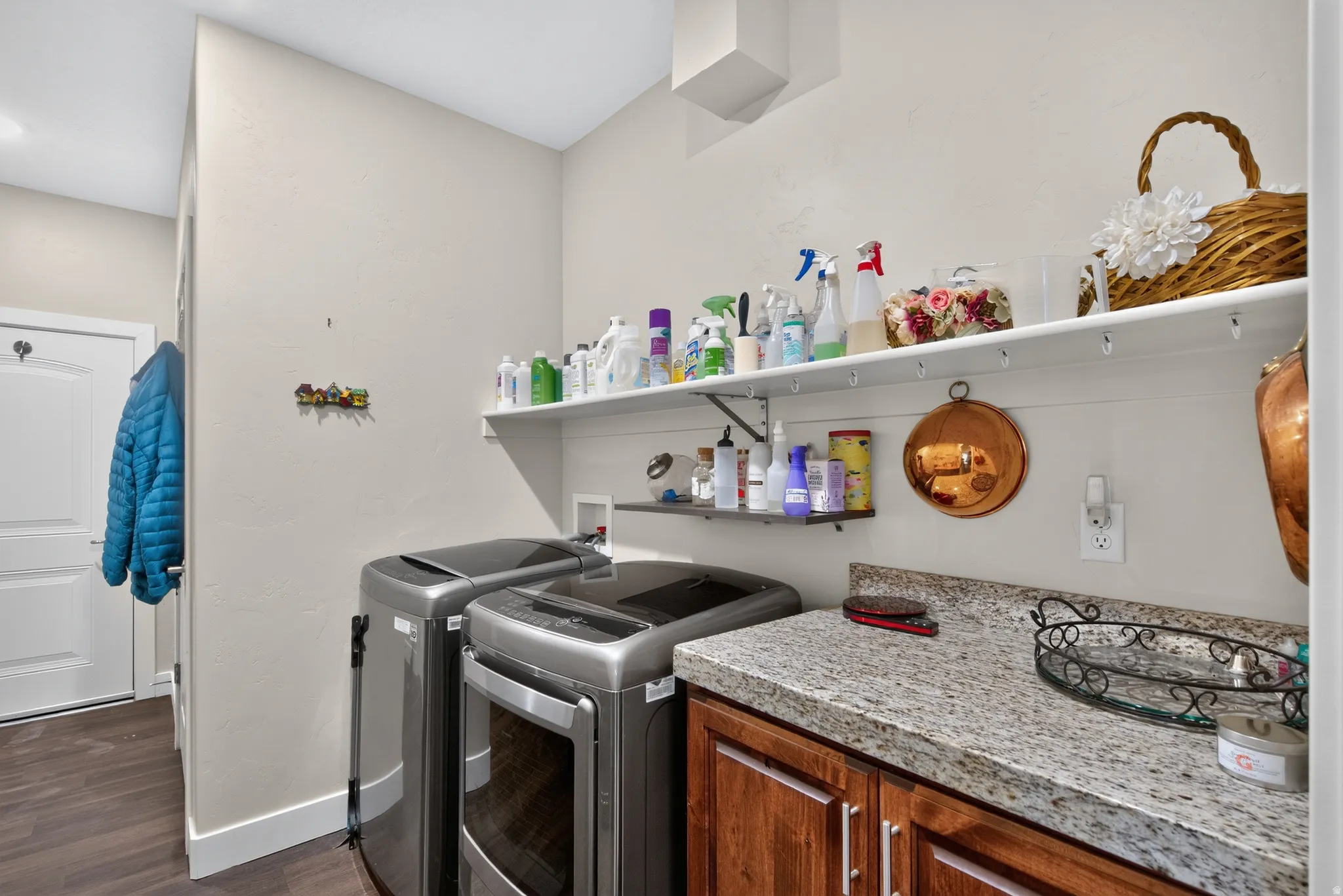 Efficient laundry room featuring washer and dryer unit, granite-style countertop with ample workspace, open shelving for storage and organization, wood-finish cabinetry, and hard-surface flooring throughout