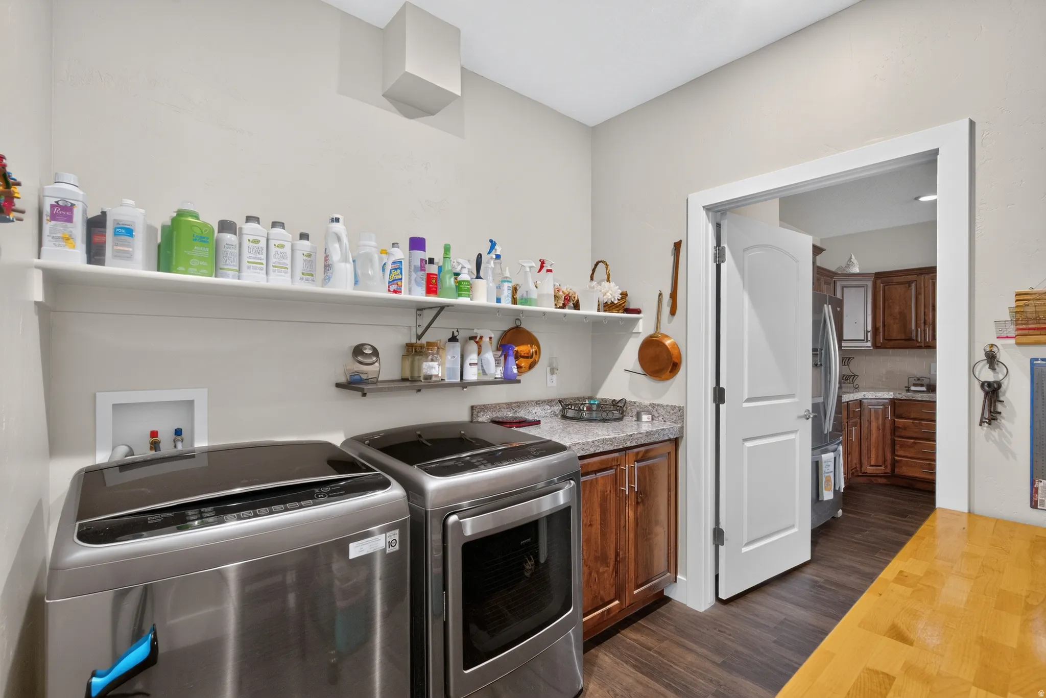 Laundry room featuring stainless steel front-load washer and dryer, wood-finish cabinetry, floating shelving with ample storage for household essentials, and direct access to the kitchen via white interior door