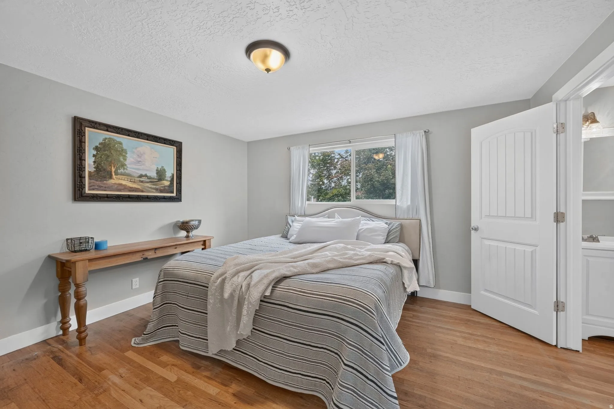 Bright secondary bedroom showcasing hardwood flooring, a metal-frame bed with neutral striped bedding, large double-hung windows with vertical blinds, white six-panel closet doors, and a wooden desk with shelf storage