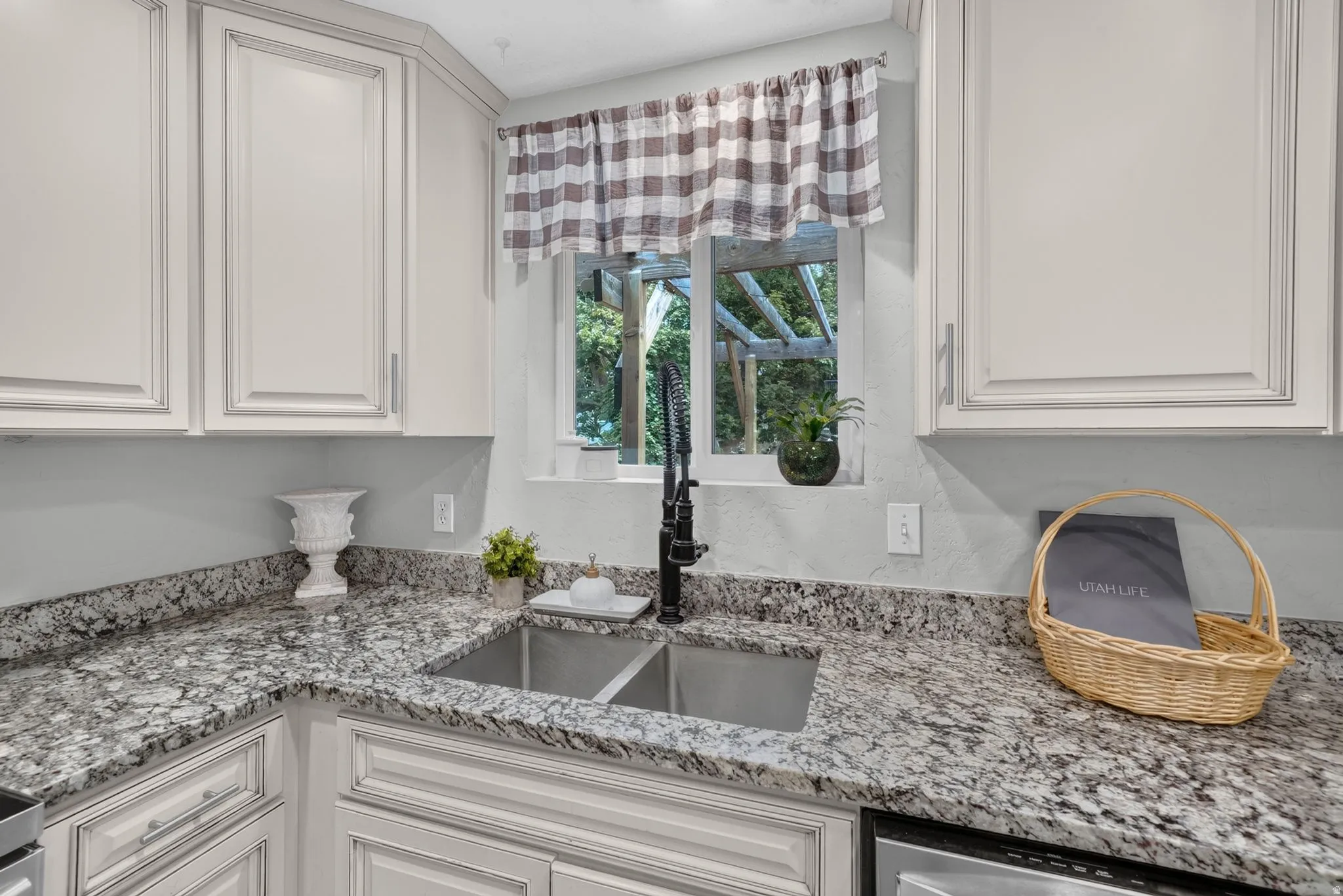 Elegant kitchen featuring white raised-panel cabinetry, speckled stone countertops with waterfall edge, stainless steel undermount sink, and a multi-pane window with gingham valance overlooking mature landscaping