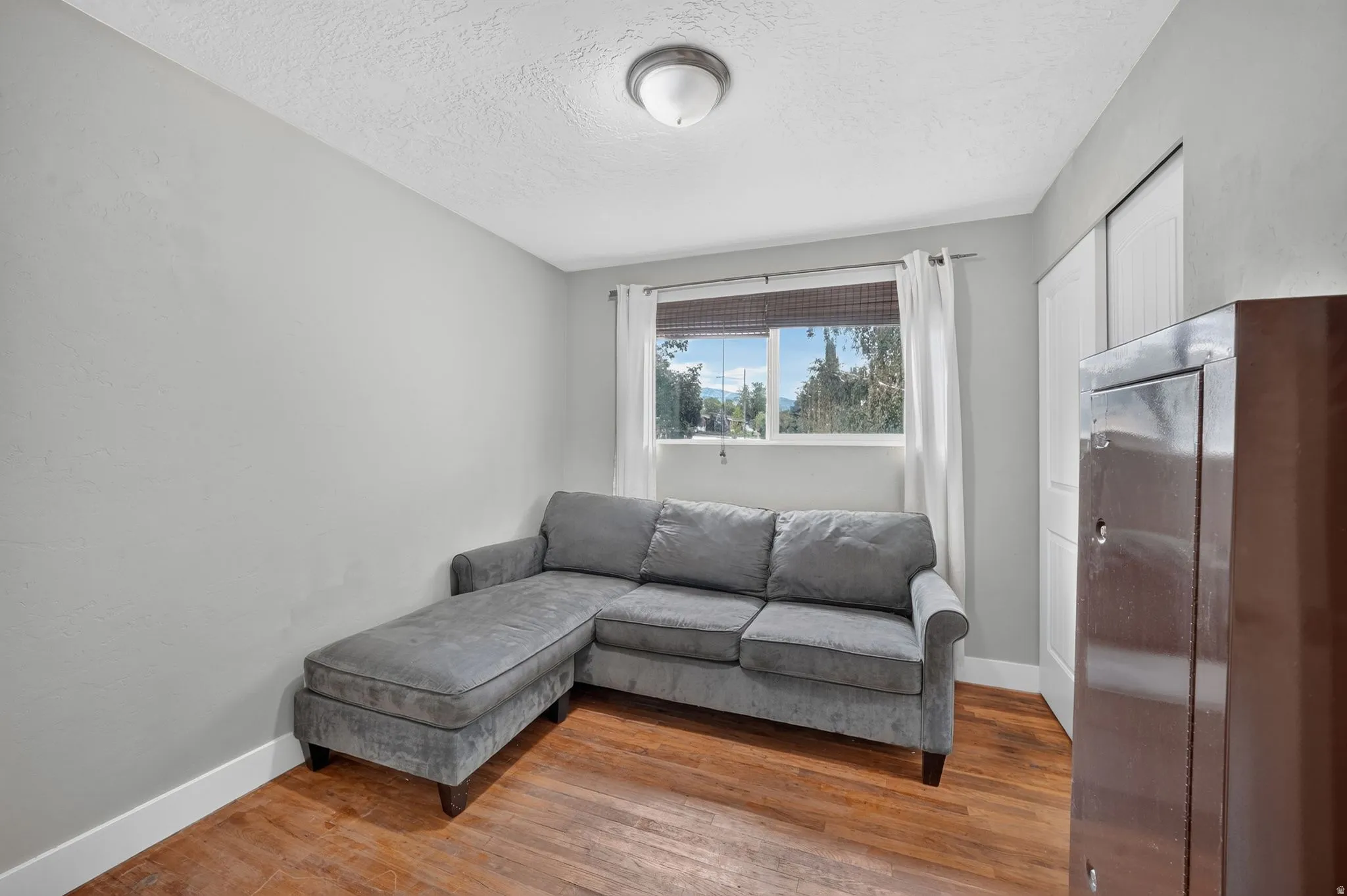 Bright corner room featuring warm wood-finish flooring, large window with natural light, and a gray upholstered sectional sofa