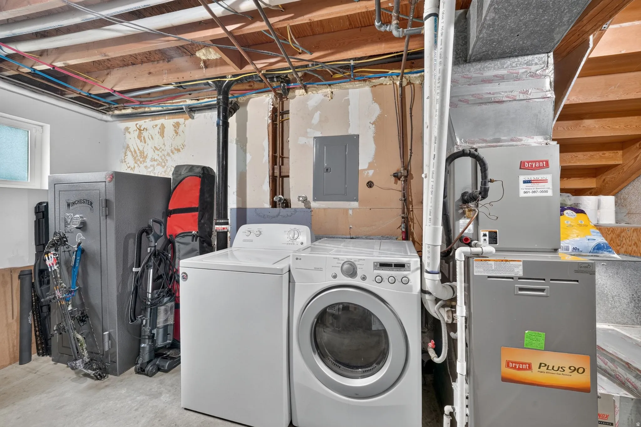 Utility room featuring full-size front-load washer and dryer, gray cabinetry with orange accents, exposed ceiling with mechanical systems, concrete flooring, and organized wall-mounted storage for household equipment and supplies