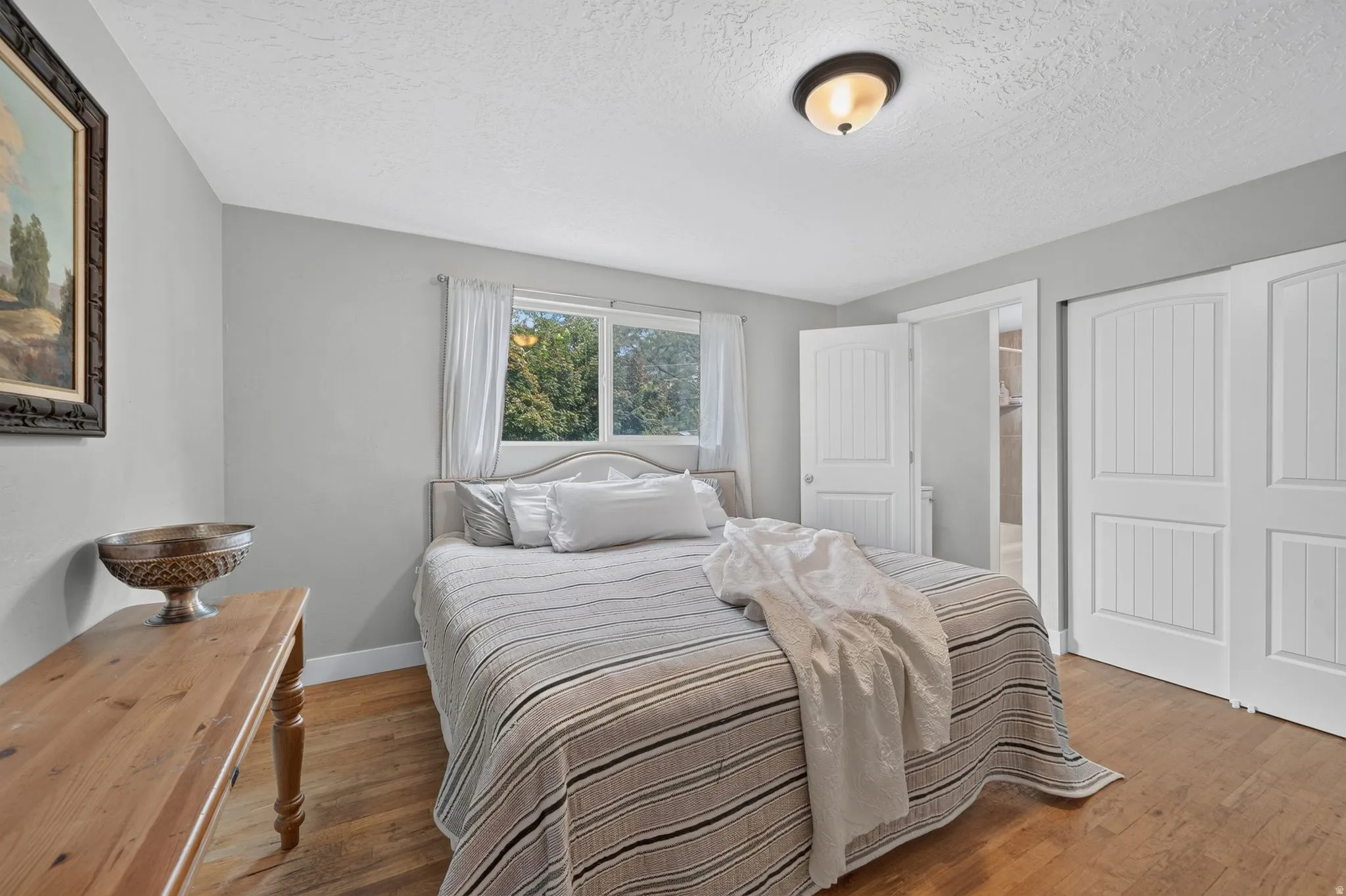 Bright secondary bedroom showcasing hardwood flooring, large double-hung windows with natural light, white six-panel closet doors, and a wood dresser