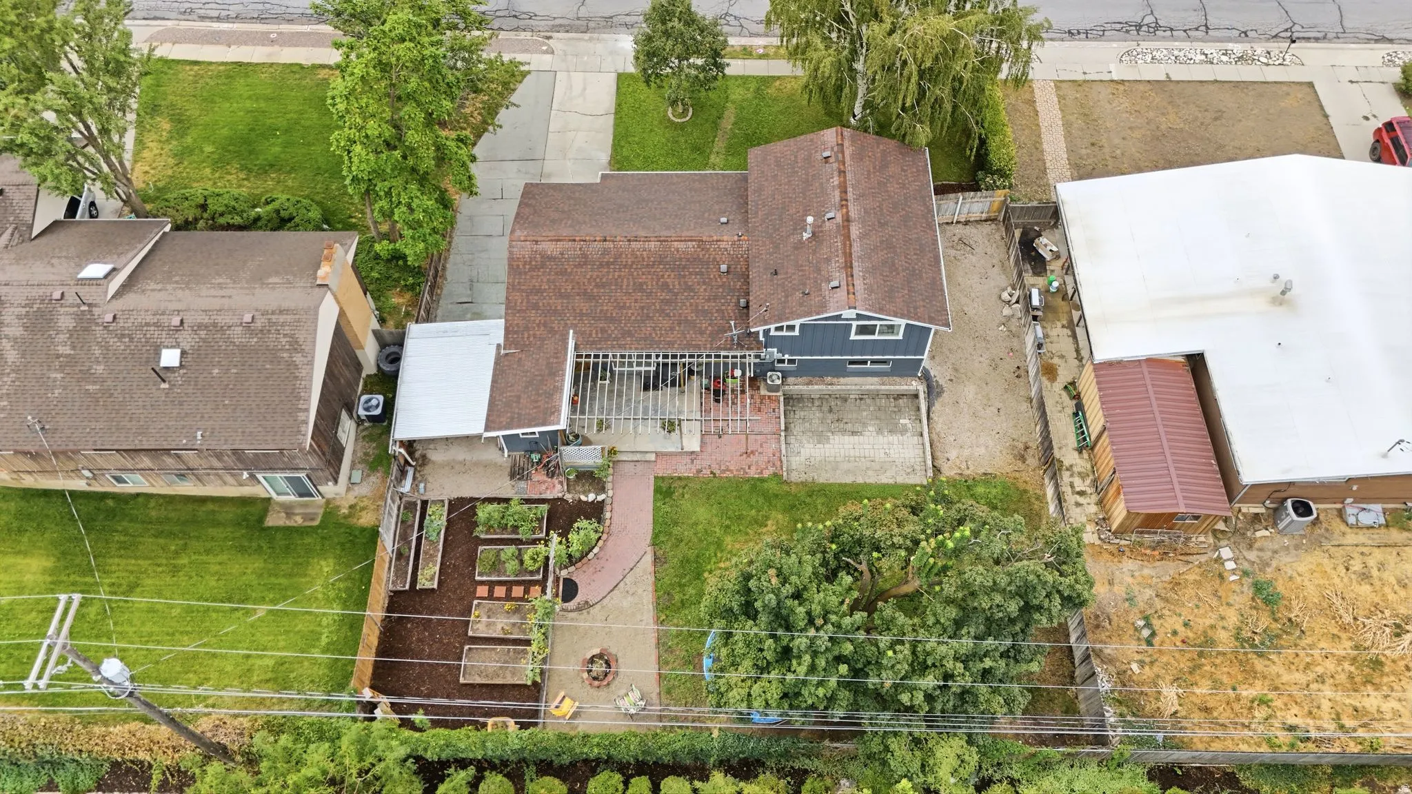 Aerial overview of the property showcasing a well-appointed residential lot with multiple structures, mature landscaping including established trees and shrubs, manicured lawn areas, brick and metal roofing, and defined hardscaping with paved pathways and
