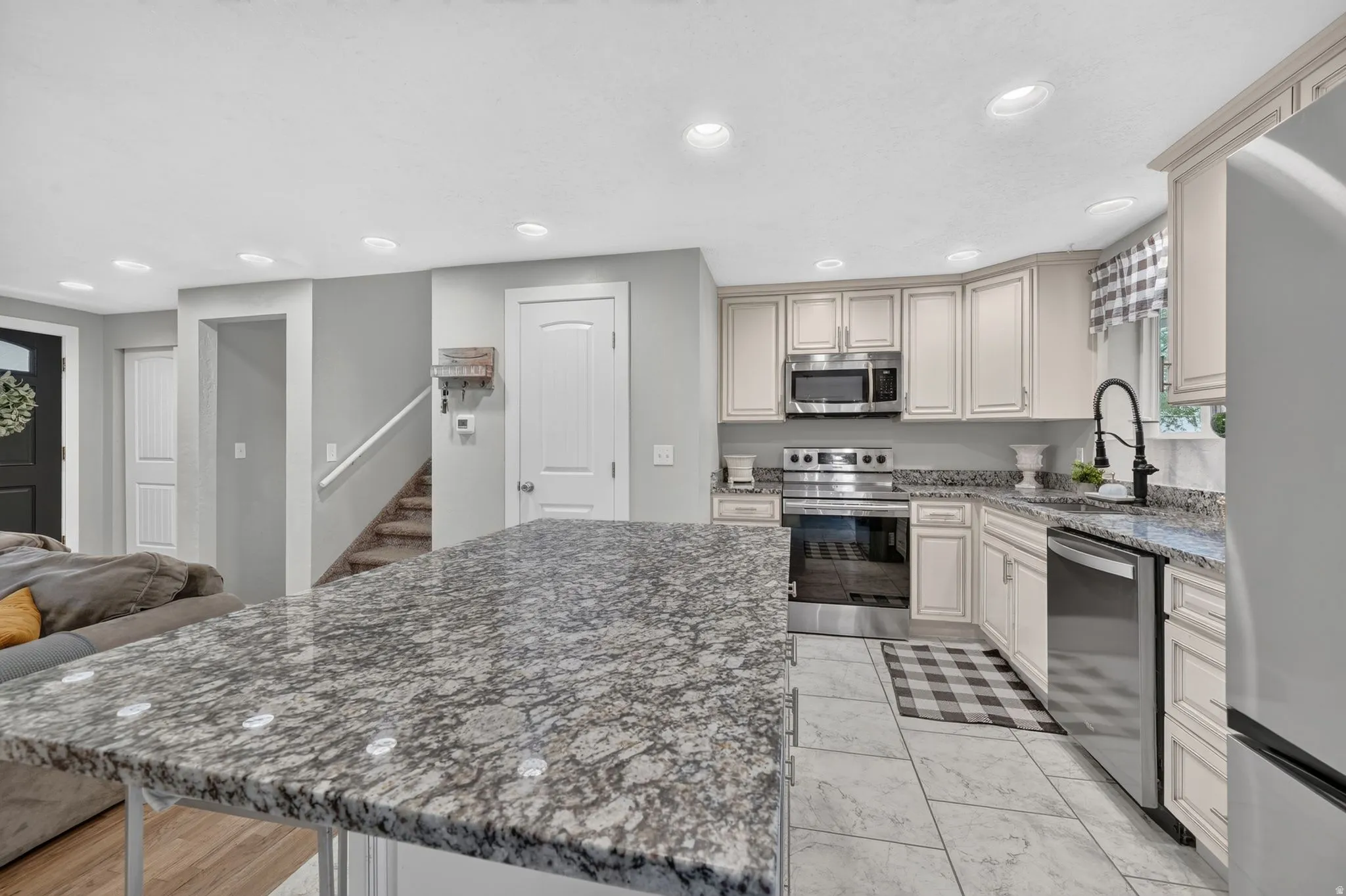 Spacious kitchen and dining area featuring cream-colored cabinetry, granite countertops, stainless steel appliances including a gas range and built-in microwave, undermount sink with pull-down faucet, recessed ceiling lighting, and neutral tile flooring