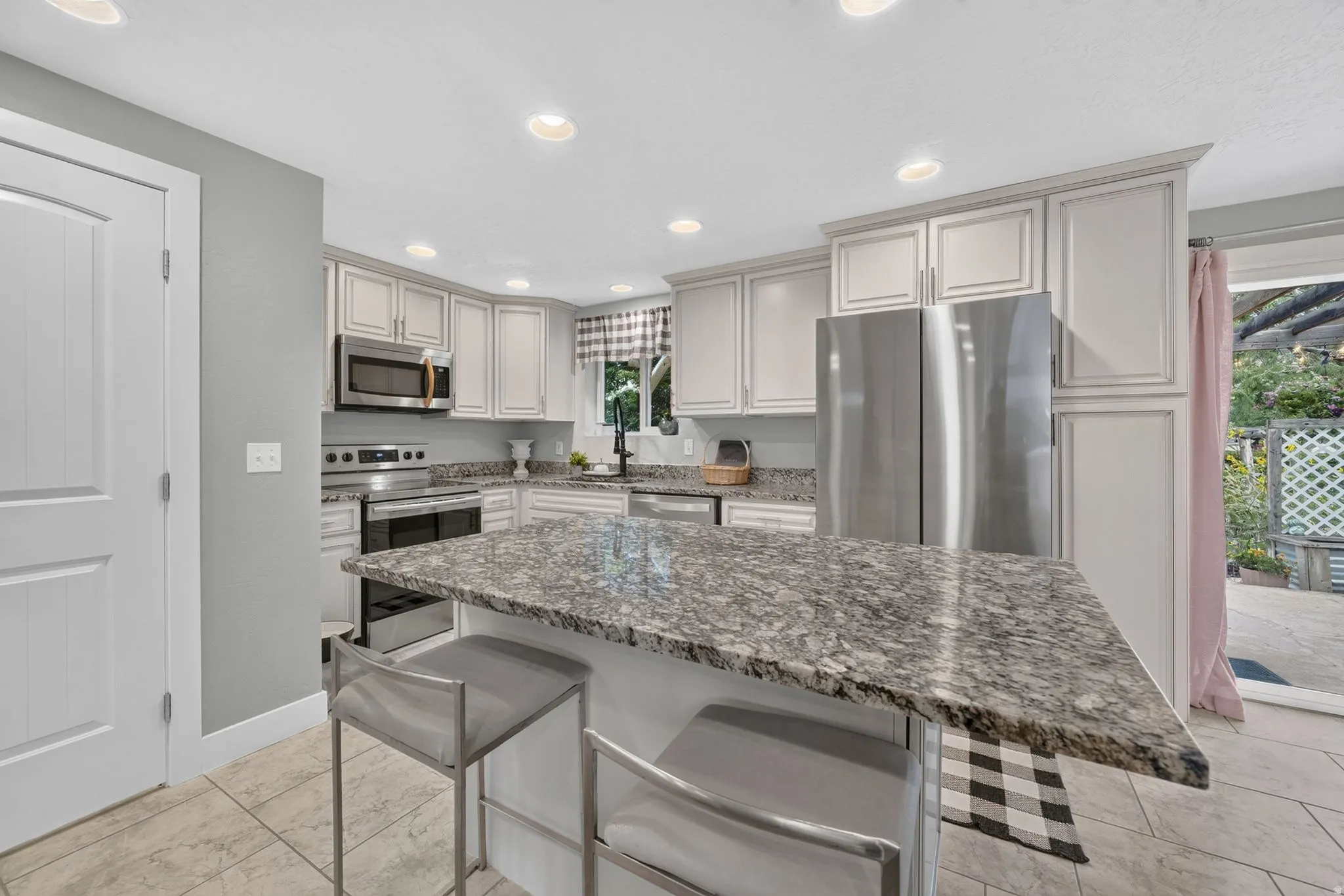 Modern kitchen featuring a spacious granite-topped island with seating, stainless steel appliances including a microwave and range, soft-close cabinetry with brushed nickel hardware, recessed ceiling lighting, and stone-finish flooring throughout the open