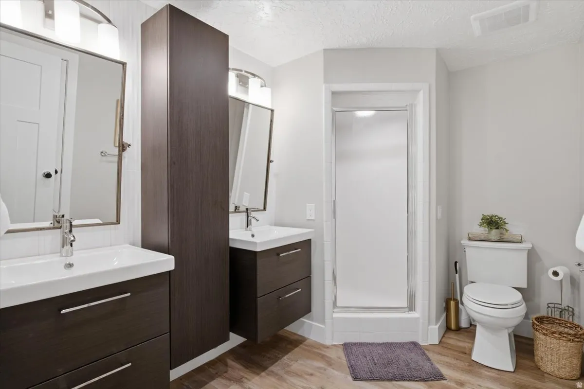 Double floating vanities with integrated sinks, wood-finish flooring, full-height storage cabinet, and a glass-enclosed shower