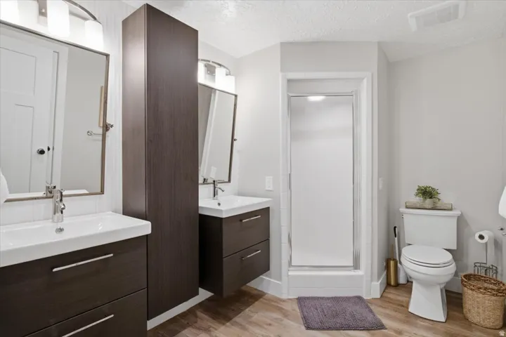 Double floating vanities with integrated sinks, wood-finish flooring, full-height storage cabinet, and a glass-enclosed shower