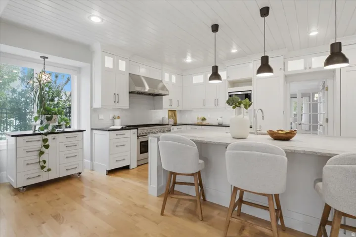 White shaker cabinetry, stainless steel range with hood, wood-finish flooring, central island with seating, and recessed and pendant lighting