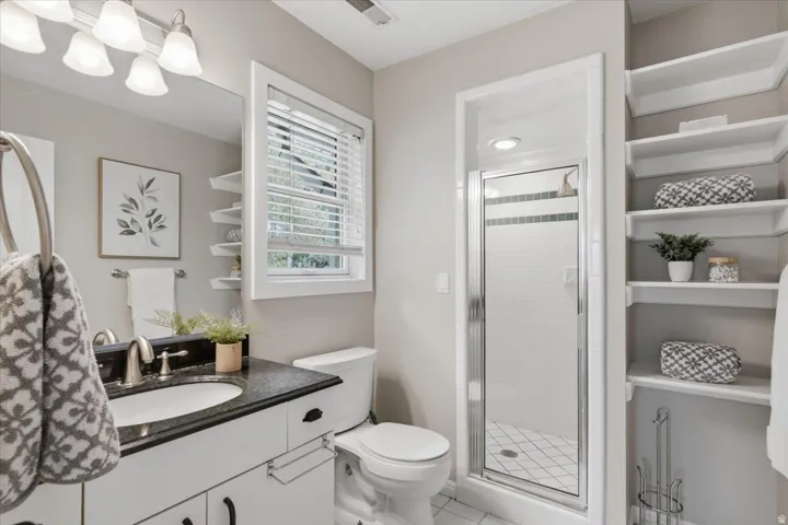 White vanity with dark countertop and undermount sink, mirrored wall, white window blinds, and built-in shelving