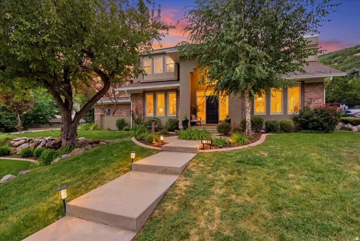 Stucco and brick facade with multiple windows, mature trees, landscaped planting beds, and a concrete pathway with integrated lighting