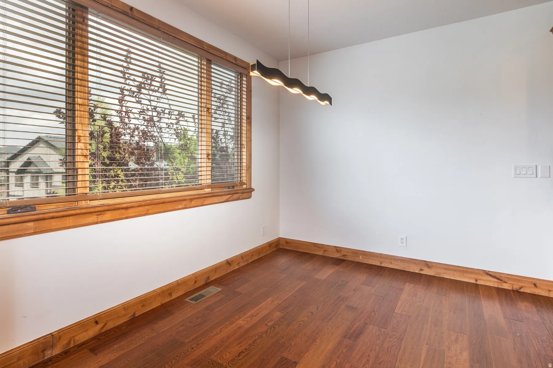 Bright secondary bedroom showcasing warm wood-finish flooring, white walls, and horizontal wood-slat window treatments framing natural light from an oversized window