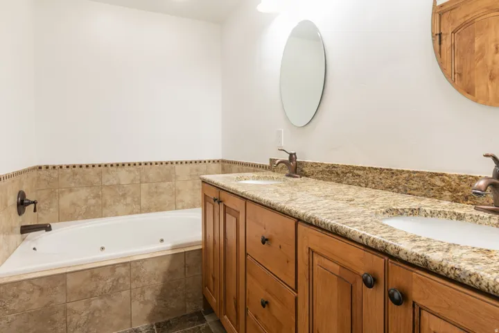 Spa-inspired master bathroom featuring a soaking tub with tile surround, dual-sink vanity with warm wood cabinetry and granite countertops, oval mirror, and brushed nickel fixtures
