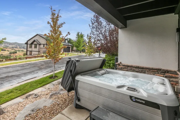 Covered patio with built-in hot tub featuring jets and integrated controls, positioned to overlook manicured landscaping with mature trees and rolling hillside backdrop