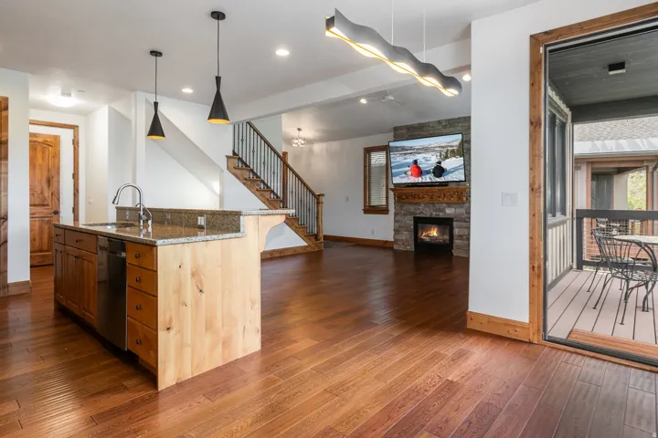 Open-concept living space featuring warm wood-finish flooring, custom wood-trimmed island with undermount sink, stainless steel pendant lighting, stone fireplace accent wall with built-in media nook, and direct access to exterior deck through glass slidin