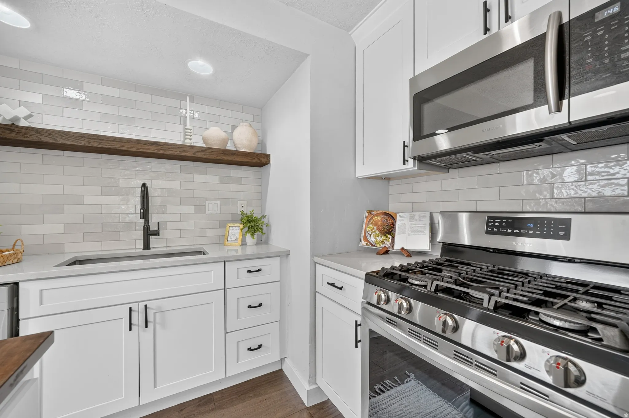 White shaker cabinetry, light-toned countertops, stainless steel appliances, white subway tile backsplash, and a dark wood-finish floating shelf