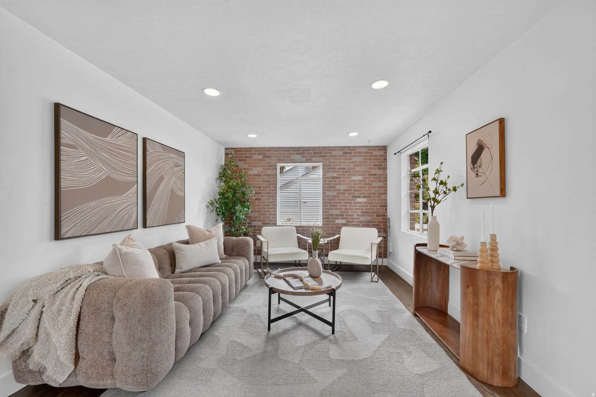 Sophisticated living room showcasing exposed brick accent wall, recessed ceiling lighting, hardwood cabinetry with wood-finish surfaces, neutral wood-finish flooring, and large windows providing natural illumination throughout the space