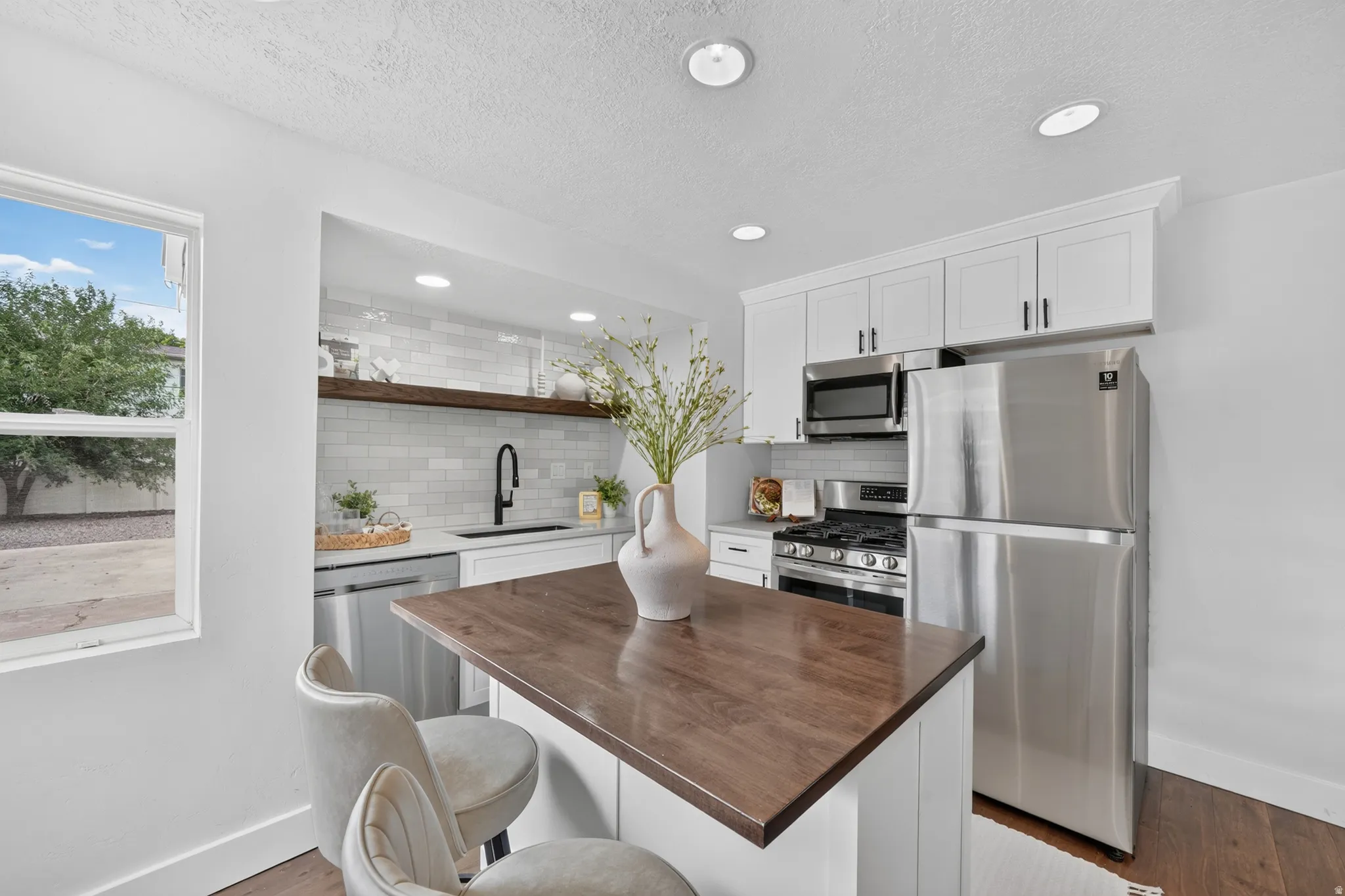 Modern kitchen featuring stainless steel appliances, white cabinetry with hardware accents, wood-finish countertop island with seating, subway tile backsplash, recessed ceiling lighting, and large windows providing natural light and outdoor views