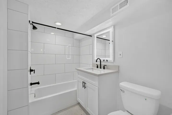 White subway tile shower surround, matte black fixtures, white vanity with stone countertop, round vessel sink, and illuminated mirror