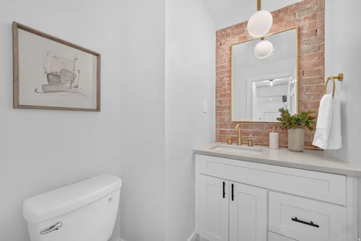 Elegant powder room featuring a white vanity with black hardware and gray countertop, exposed brick accent wall, framed mirror with integrated lighting, and white ceramic toilet