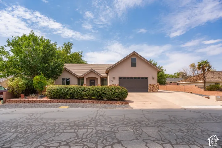 Charming stucco residence featuring a two-car garage with dark paneling, mature landscaping with established trees and hedging, brick hardscaping along the front perimeter, arched entryway detail, and a spacious driveway with stamped concrete finish