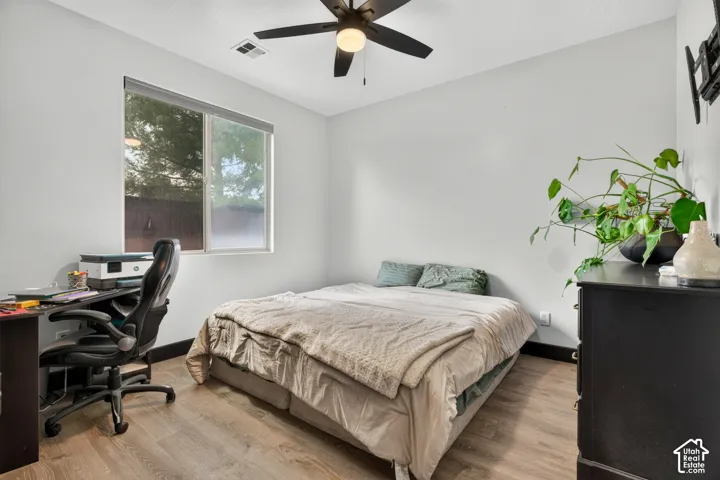 Bright secondary bedroom with wood-finish flooring, large dual-pane windows providing natural light, ceiling fan with integrated lighting, and dedicated workspace with built-in desk and ergonomic seating
