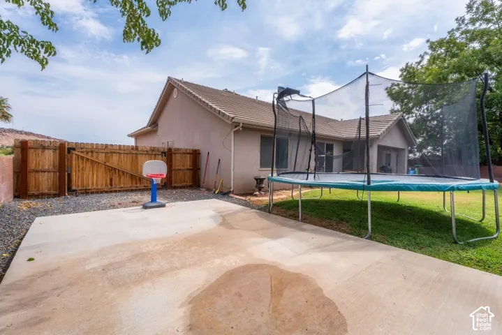 Spacious backyard oasis featuring a concrete patio, regulation basketball hoop, trampoline with safety enclosure, mature shade trees, and manicured lawn