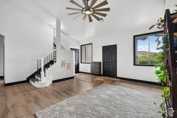Spacious open-concept living area featuring wide-plank wood-finish flooring, soaring white ceilings with modern windmill-style ceiling fan, and abundant natural light from large windows