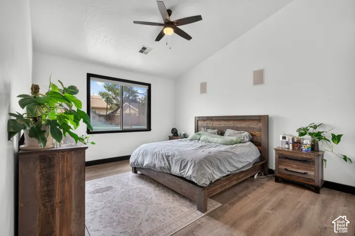 Primary bedroom suite featuring a wood-finish platform bed frame, wood-finish nightstand with drawer storage, ceiling fan with integrated lighting, large fixed window with black frame, and wood-finish flooring throughout