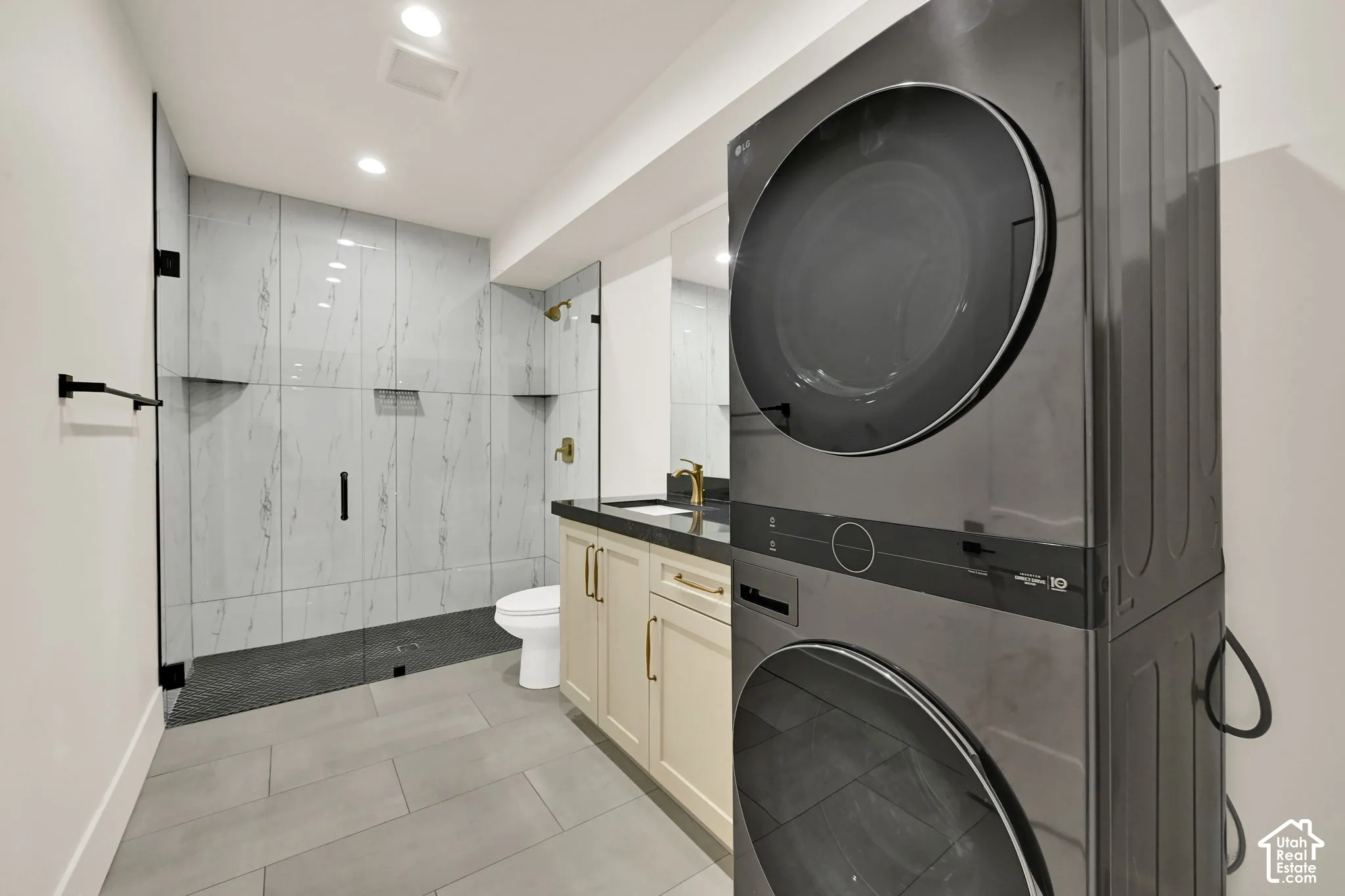 Built-in stacked washer and dryer, glass-enclosed shower with black fixtures, vanity with gold hardware, recessed lighting, and tile flooring