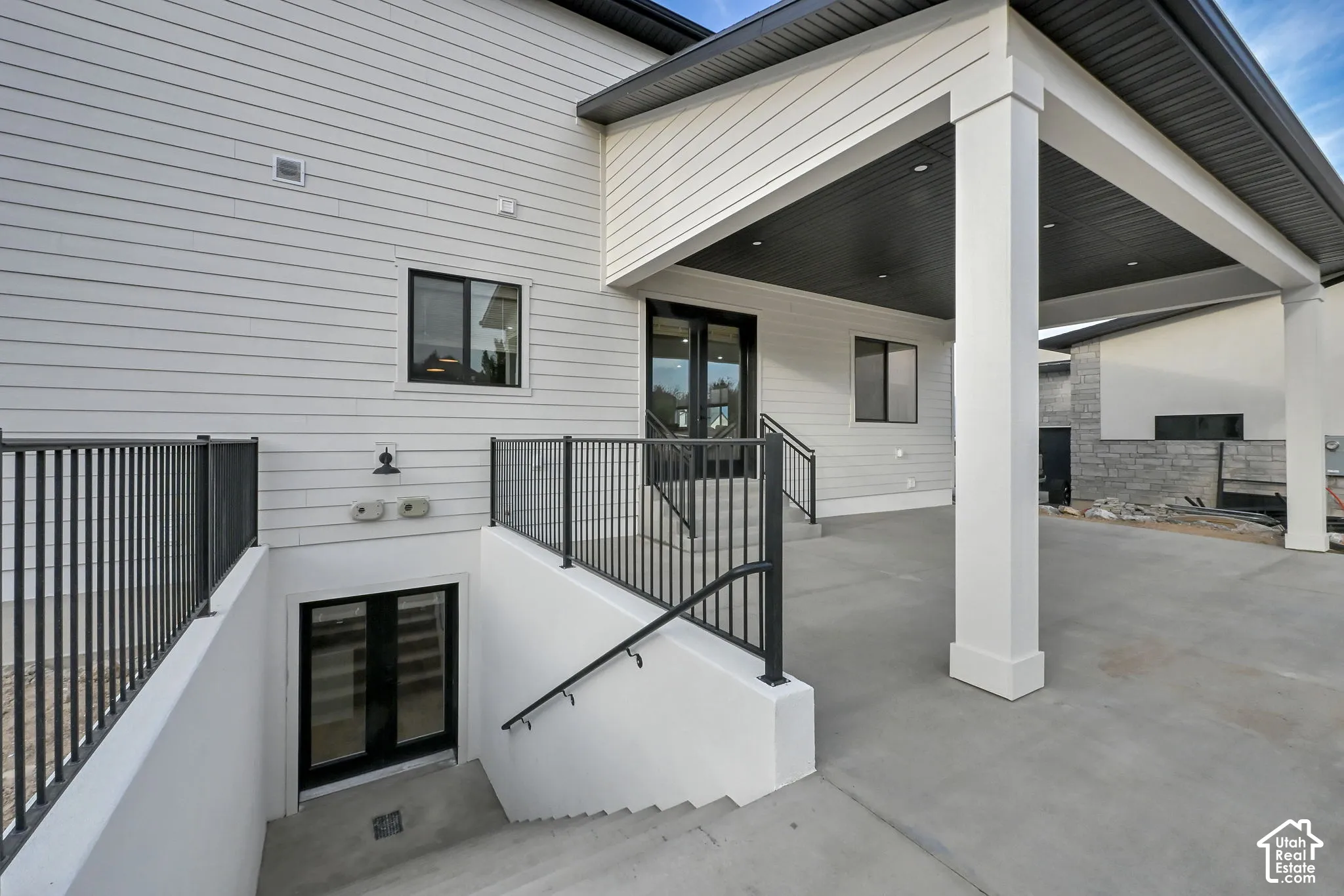 Covered concrete patio with recessed lighting, white columns, black railings, and a walk-out basement entrance