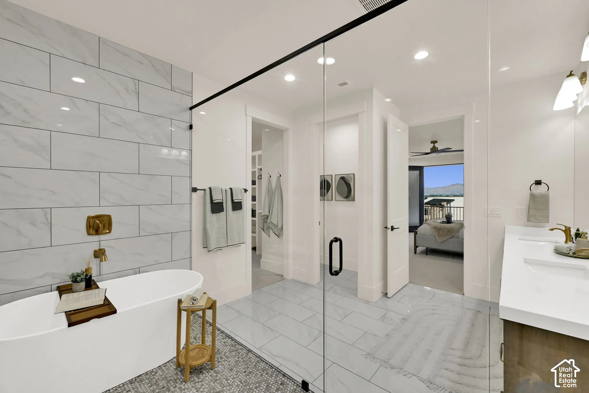 Freestanding soaking tub with a wall-mounted brass faucet, glass-enclosed shower, white tile flooring, and a double vanity with undermount sinks