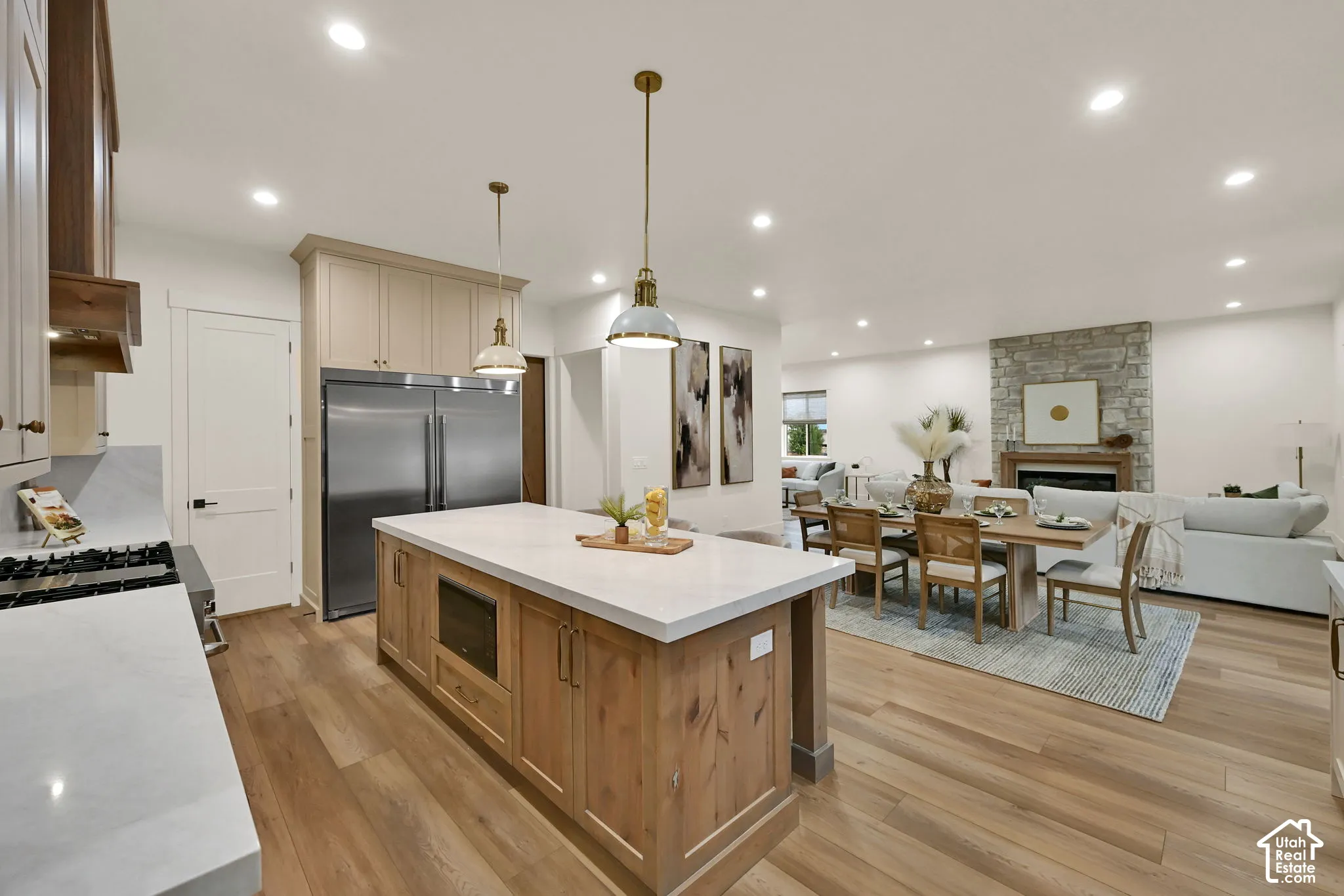 Open-concept living area with wood-finish flooring, a stone-faced fireplace, recessed lighting, and a kitchen island with a light-toned countertop