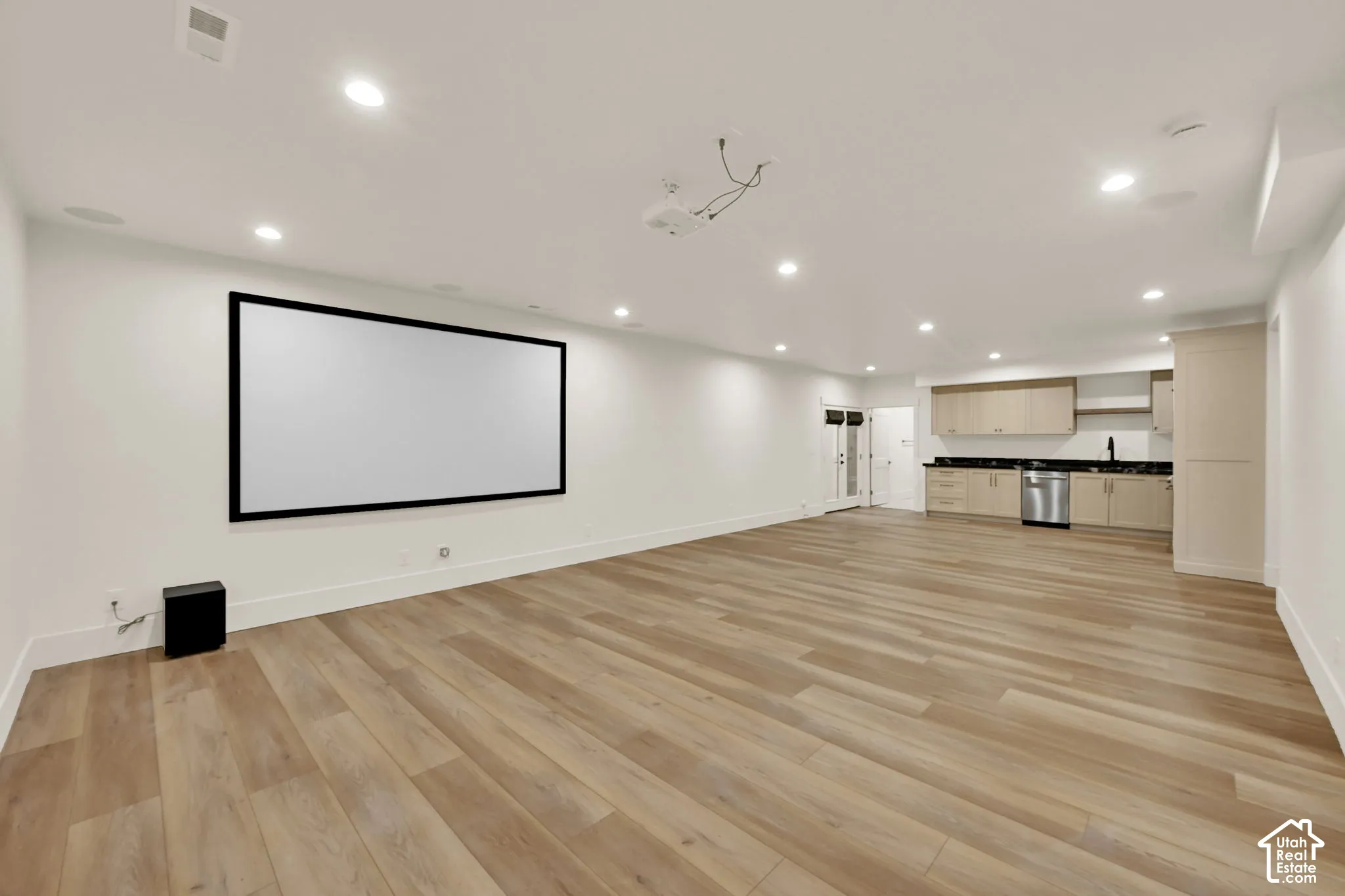 Spacious entertainment area featuring a large projection screen, recessed lighting, wood-finish flooring, and a compact kitchen with a stainless steel dishwasher