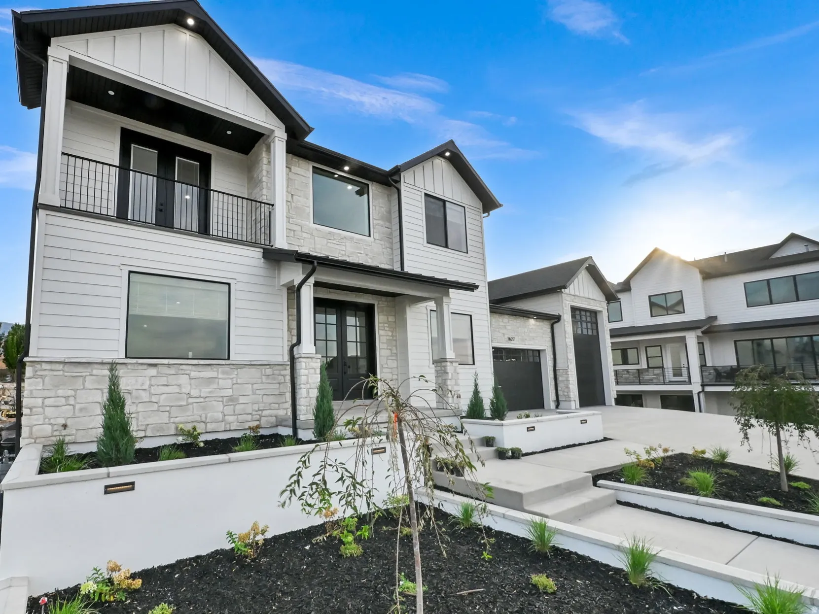 Modern exterior featuring white siding, stone accents, black window frames, a second-story balcony with metal railing, and a covered entry